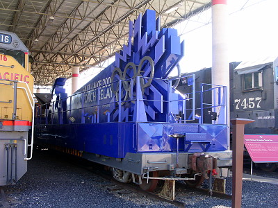 [A flat railcar with blue sides and a blue snowflake atop a torch which is on its side resting in the car. There are railcars on either side of this blue one under a large roof.]